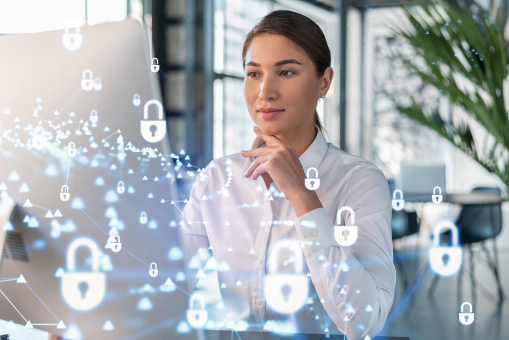 Attractive businesswoman in white shirt at workplace working with laptop to defend customer cyber security. Concept of clients information protection. Padlock hologram over office background.