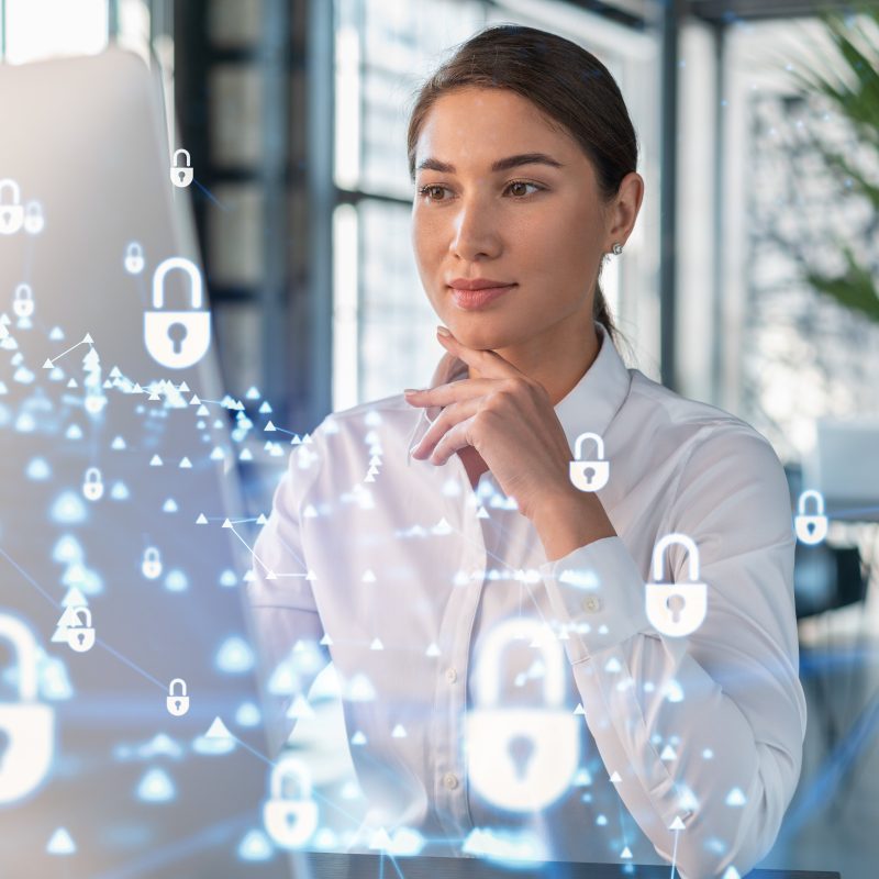 Attractive businesswoman in white shirt at workplace working with laptop to defend customer cyber security. Concept of clients information protection. Padlock hologram over office background.